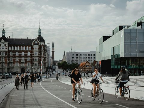 two people biking and other people walking on pathway near buildings under white and blue sky