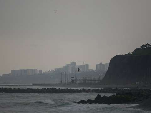 A paraglider above a coastal city skyline with rocky cliffs and ocean waves.