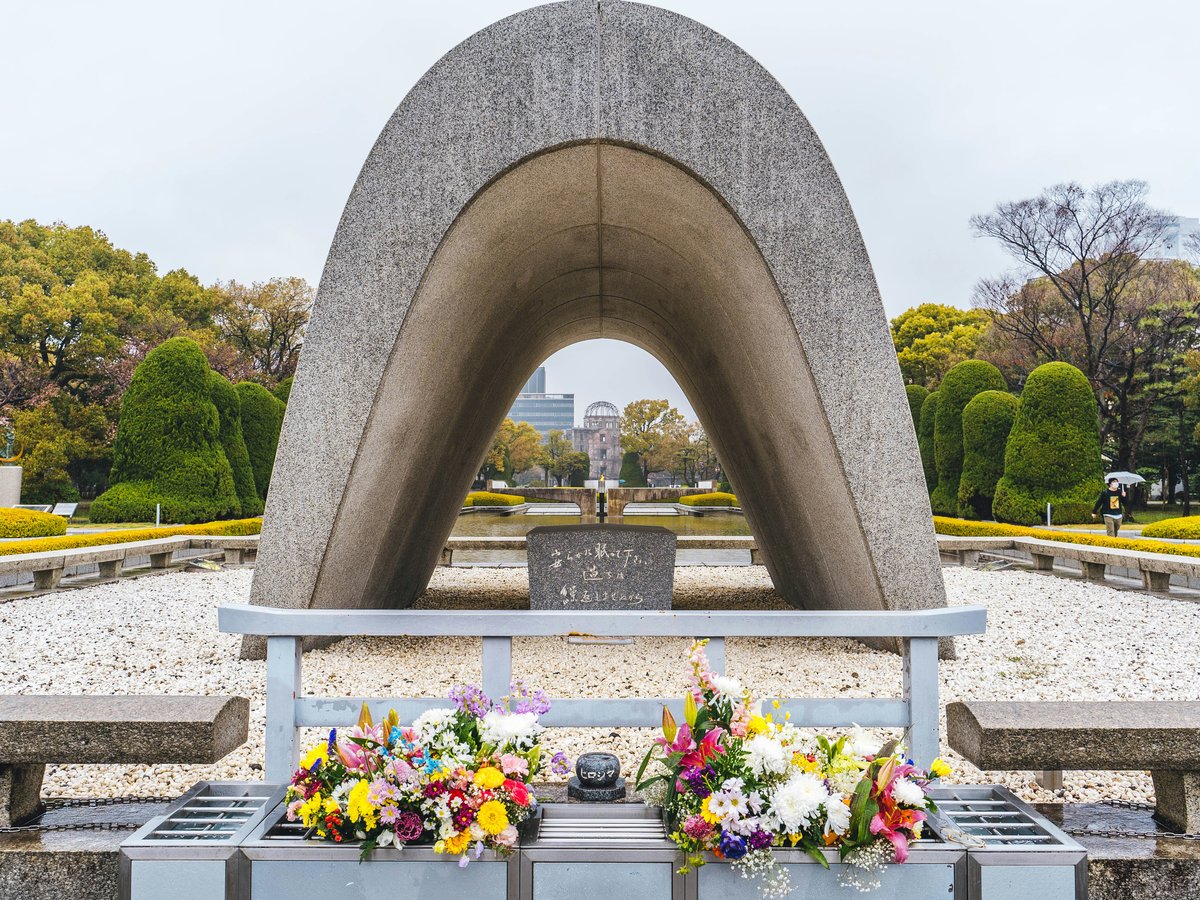 a large stone structure with flowers in front of it with Hiroshima Peace Memorial in the background