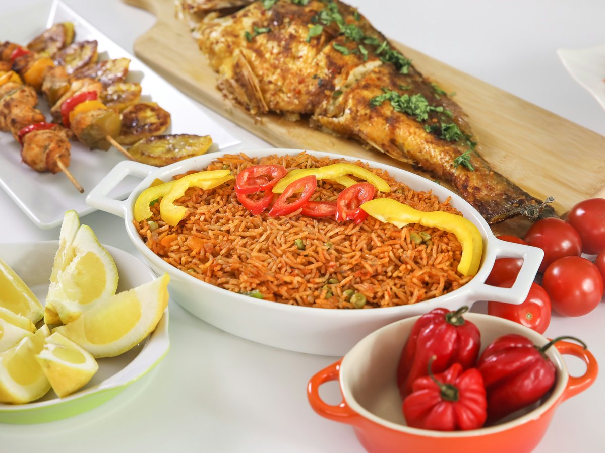 Jollof rice in a white dish with grilled fish, skewers, and fresh vegetables