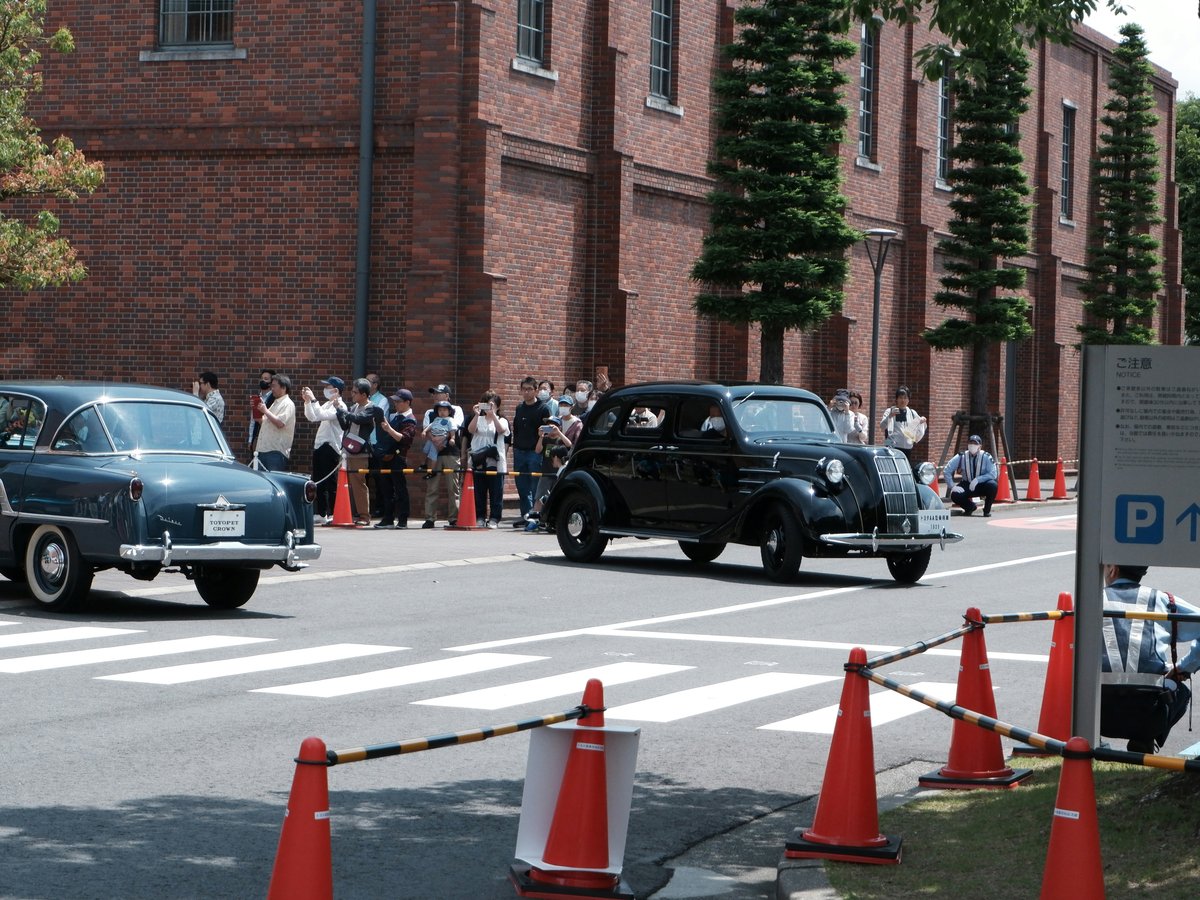 Two vintage cars drive past onlookers near brick building