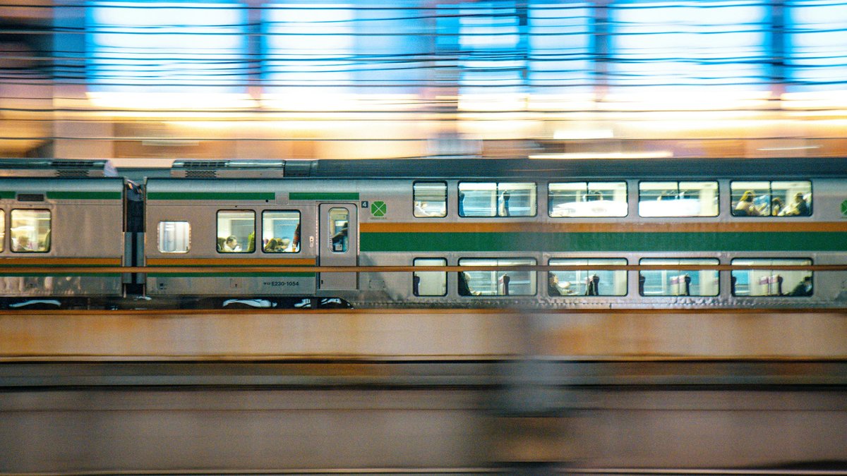 A commuter train streaks past an elevated Tokyo platform in a long-exposure blur of headlight trails, the surrounding skyline reduced to soft warm bokeh dots