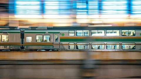 A commuter train streaks past an elevated Tokyo platform in a long-exposure blur of headlight trails, the surrounding skyline reduced to soft warm bokeh dots