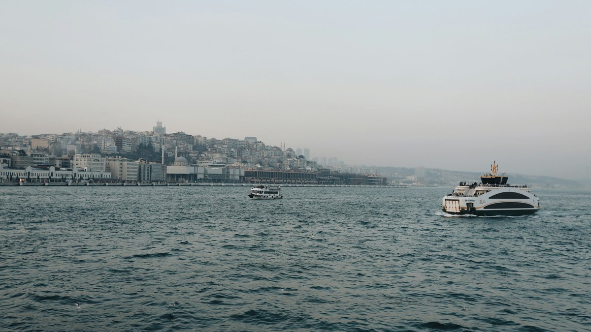 A passenger ferry crossing the calm teal waters of the Bosphorus, the domes and minarets of Istanbul's old city hazing into a muted pastel afternoon
