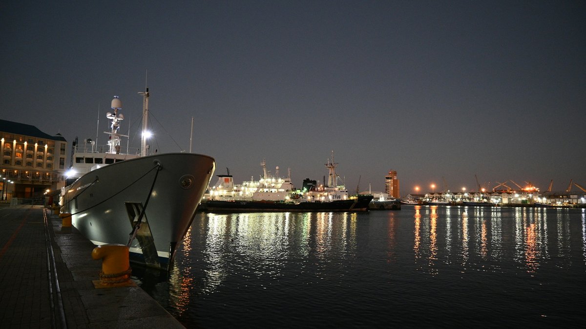 A working ship's prow looms over the V&A Waterfront quay at night, the still harbour water mirroring warm amber dock lights and distant cranes glowing against an inky charcoal sky
