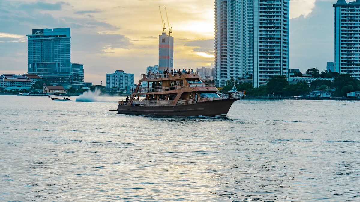 A wooden river cruiser slipping along the Chao Phraya as Bangkok's high-rise skyline catches the sunset behind it
