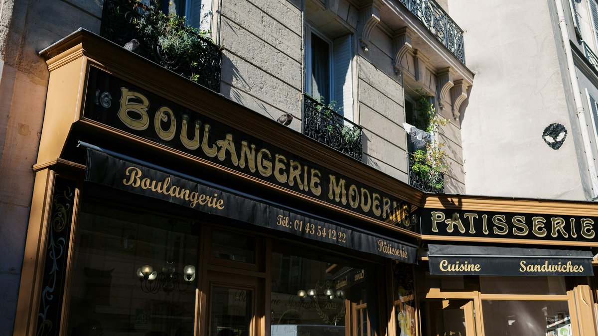 A corner Parisian boulangerie with gilded 'Boulangerie Moderne' lettering, warm storefront windows stacked beneath classic Haussmann balconies