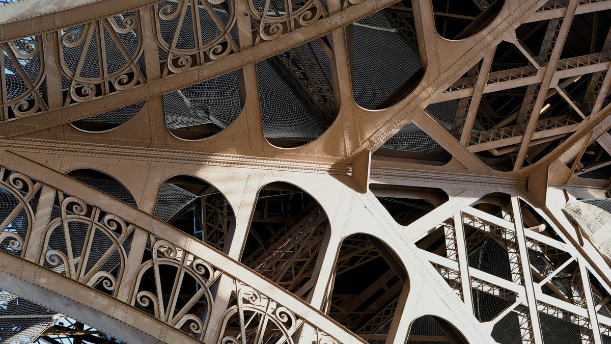 A close-up abstraction of the Eiffel Tower's cream-painted iron lattice, riveted beams fanning into geometric curves against the daylight