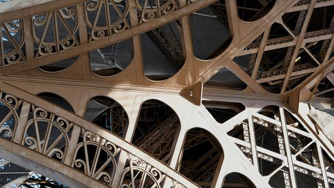 A close-up abstraction of the Eiffel Tower's cream-painted iron lattice, riveted beams fanning into geometric curves against the daylight