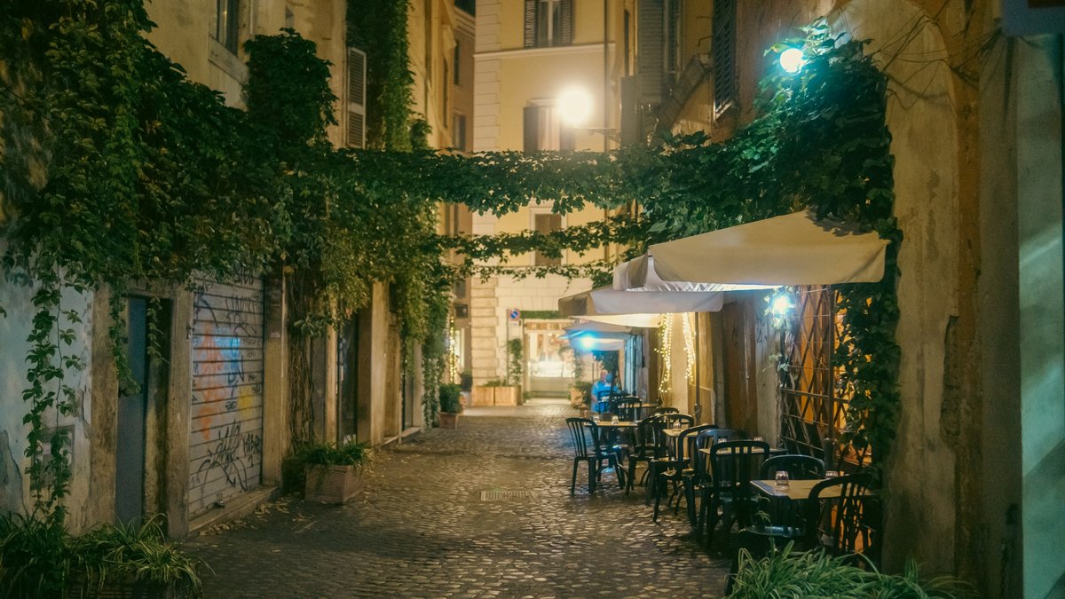 A hushed Trastevere lane after dark, cobblestones slick under warm lamplight as empty trattoria tables and ivy-draped walls recede into a soft amber glow