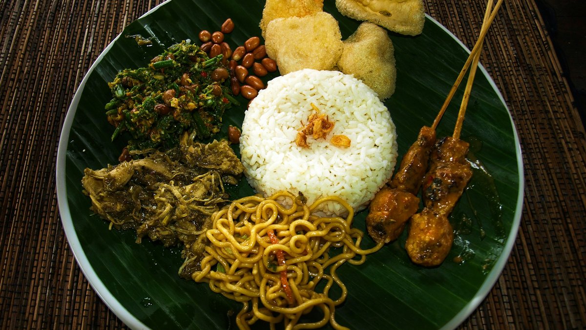 A Balinese nasi campur plate on a banana leaf — mounded steamed rice ringed by chicken satay skewers, mie goreng noodles, urab greens, peanuts, tempeh and slow-cooked jackfruit