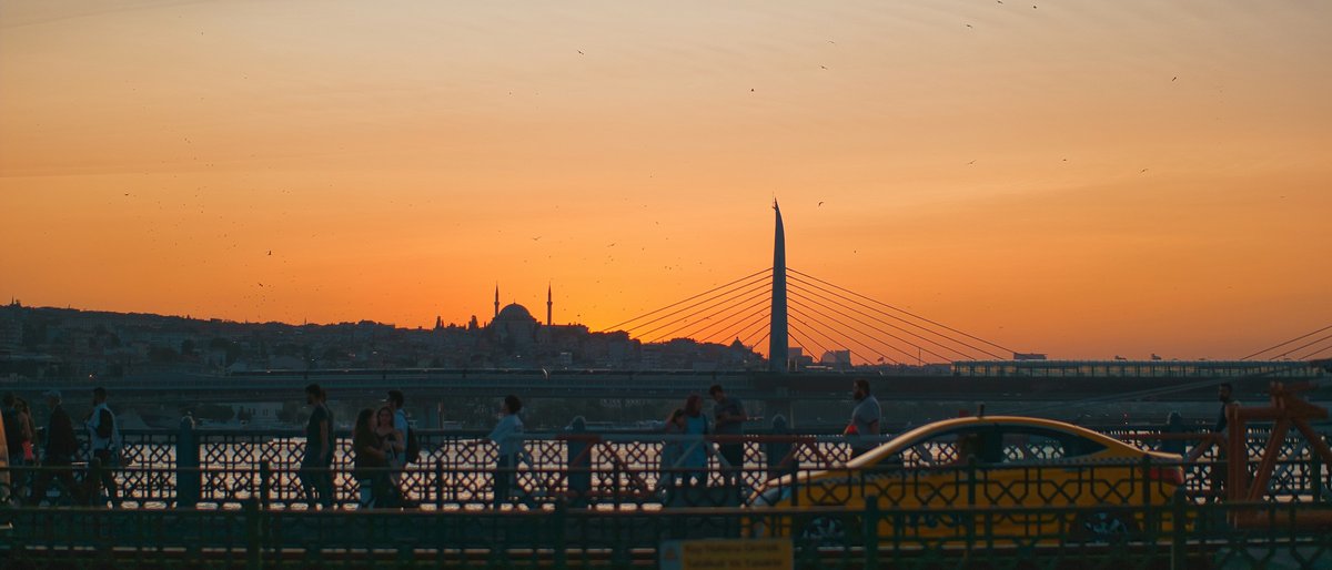 Silhouetted commuters crossing the Galata Bridge at sunset, the minarets of the old city skyline rising against a molten orange Istanbul sky
