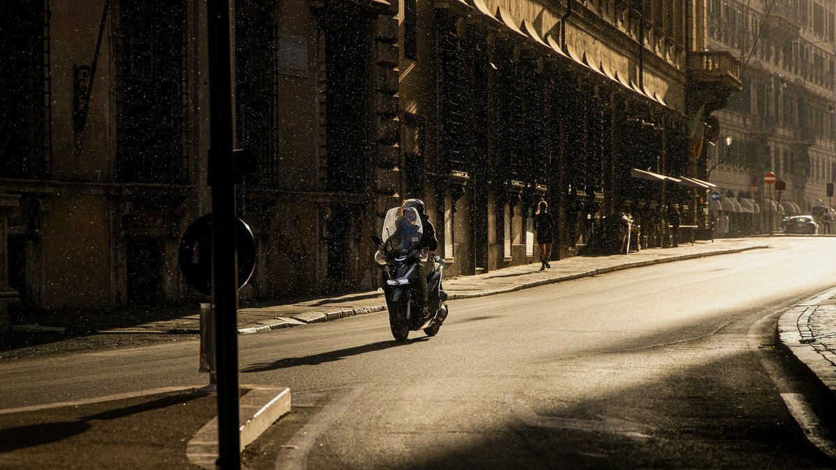 A lone rider on a scooter weaves down a rain-slicked Roman street at dusk, tall tenement facades receding into a moody violet haze lit by scattered shopfront glow