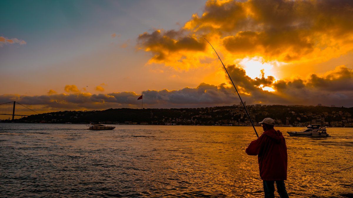 A lone fisherman silhouetted on the Bosphorus shore at golden hour, backlit clouds breaking above a passing boat on a glowing amber sea