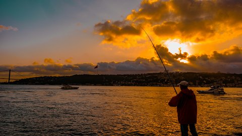 A lone fisherman silhouetted on the Bosphorus shore at golden hour, backlit clouds breaking above a passing boat on a glowing amber sea
