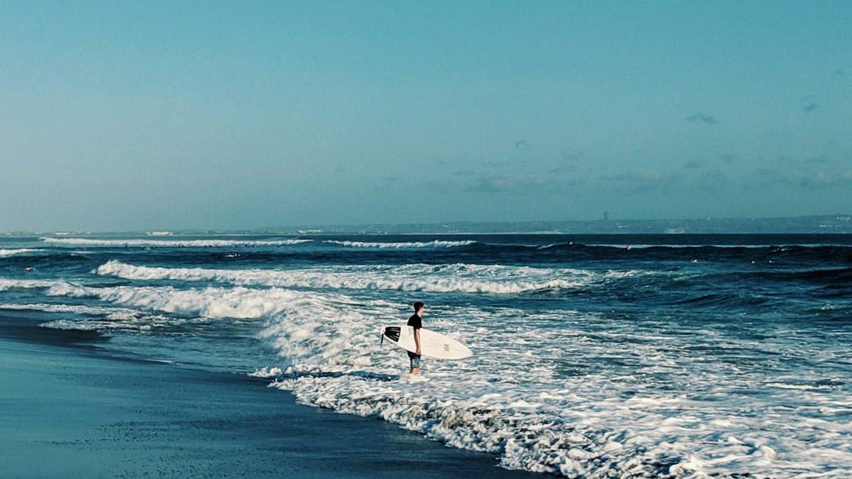 A lone surfer stands in the whitewater of a teal Bali break, board tucked under one arm, scanning offshore lines as foam curls across a long dark-sand shore at the south-coast reefs