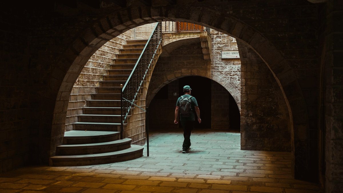 A lone backpacker framed beneath the arched stone passageway of Barcelona's Gothic Quarter, worn cobbles glowing warm amber under a single overhead lamp