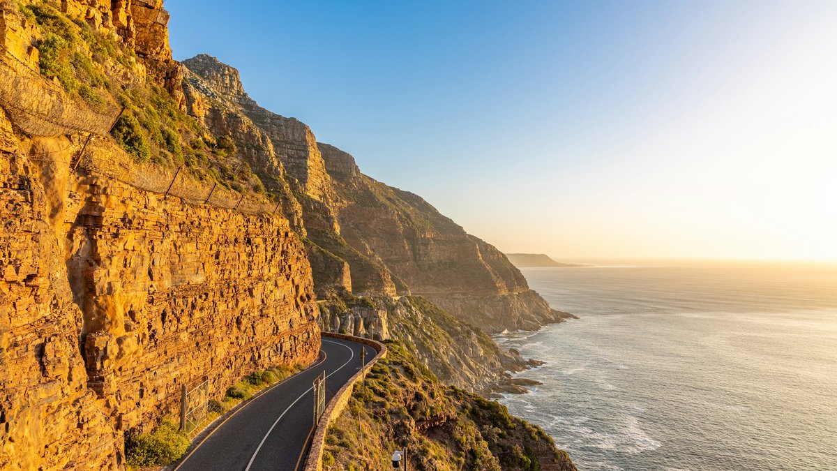 Chapman's Peak Drive curves along an ochre-gold sandstone cliff face above the Atlantic at golden hour, empty asphalt vanishing toward distant headlands as warm late light glances off rock