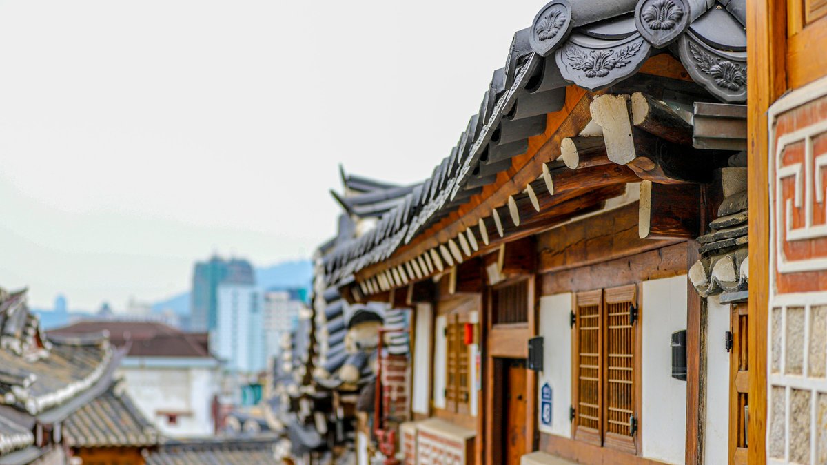 An ornate Bukchon hanok's curved eaves, carved wooden shutters and lotus-stamped tile ends frame a soft-focus sliver of modern Seoul skyline rising beyond the village rooftops