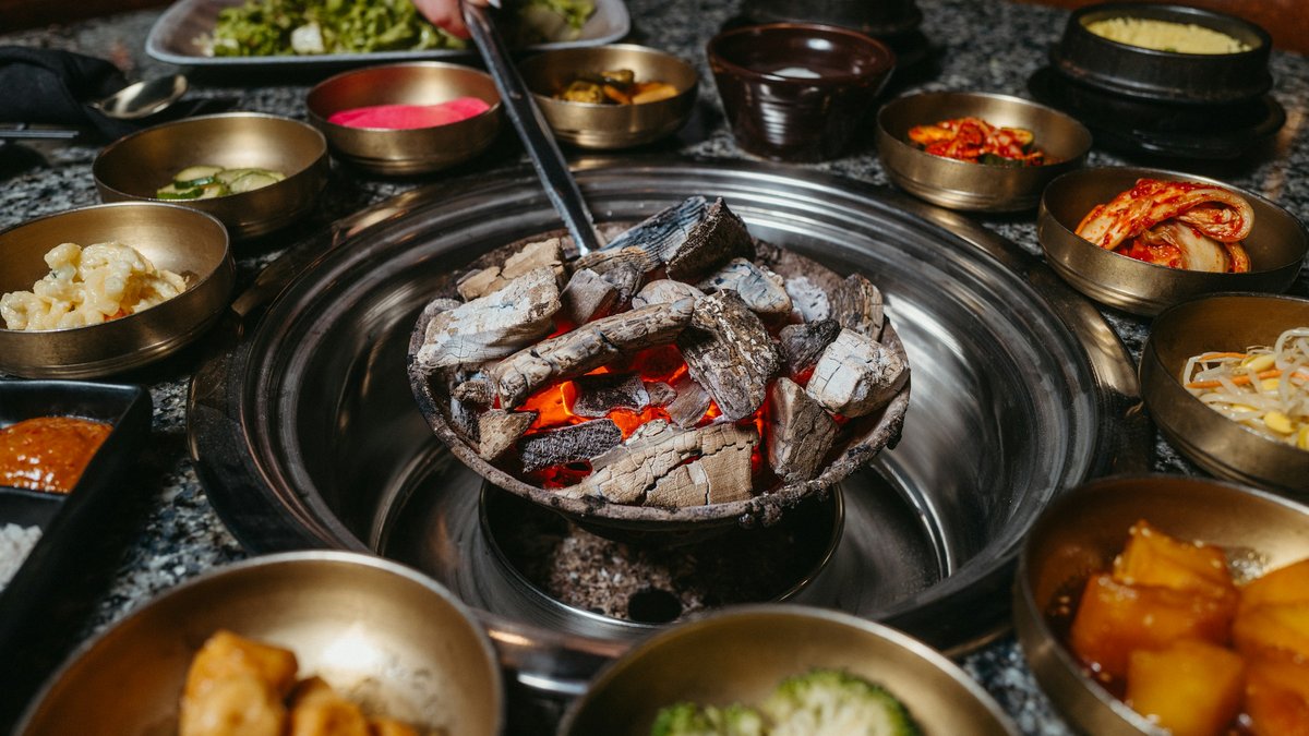 A brass ring of Korean banchan — kimchi, pickled radish, bean sprouts, braised potato — surrounds a glowing charcoal brazier waiting for the first cuts of barbecue to hit the grill