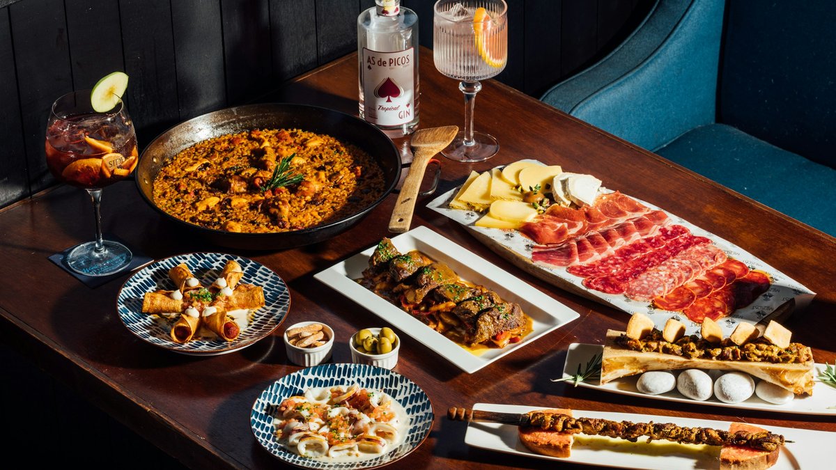 A candlelit Catalan feast: saffron-rich paella, sliced jamón and cured sausage, olives, croquetas, and a vermut spritz arranged across a dark wooden tavern table