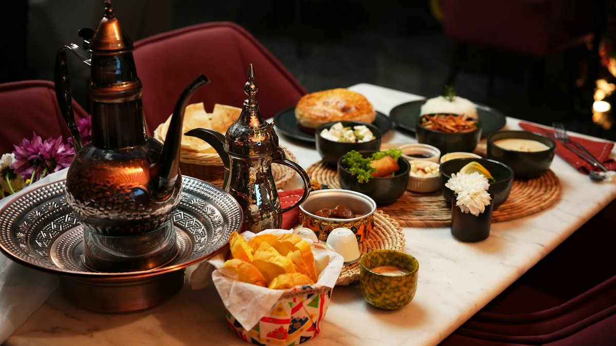 An engraved brass dallah coffee pot on an ornate silver tray anchors a dim-lit marble table laid with bowls of mezze, flatbread, luqaimat, date halwa and a basket of golden chips against deep burgundy banquettes