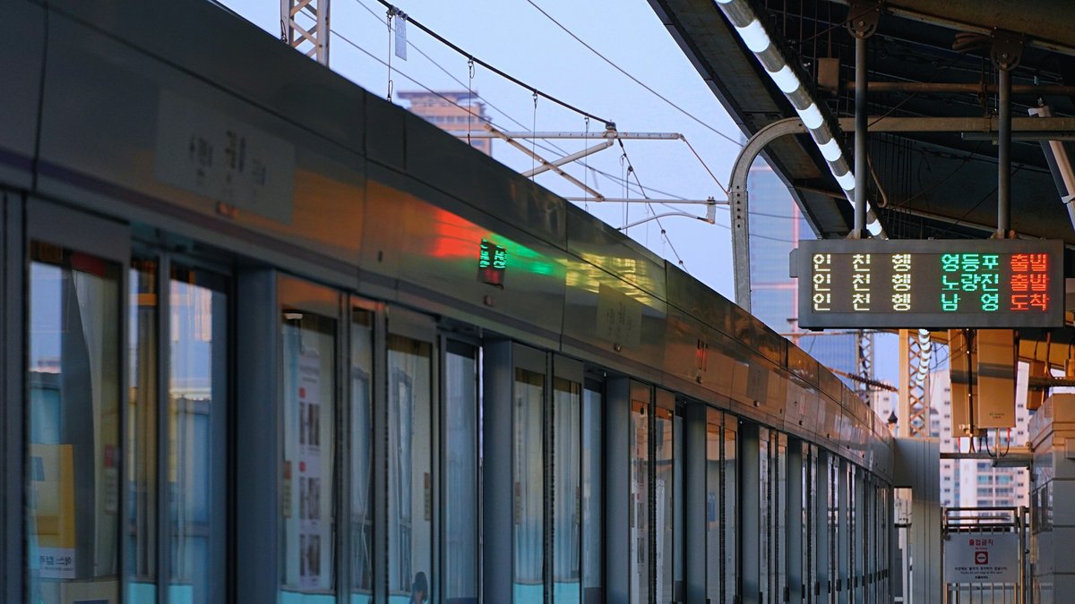 A Seoul Metro Line 1 train waits at an elevated platform, blue glass screen-doors reflecting dusk sky as an orange Hangul destination board glows with stops along the Incheon run
