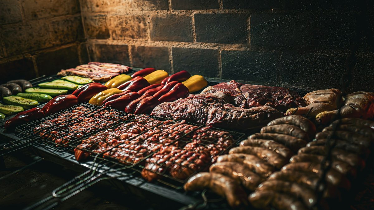A full Argentine asado laid across the parrilla grate — coiled chorizos and morcillas, chinchulines, short ribs, sliced red and yellow bell peppers — lit by warm embers against a soot-blackened brick wall