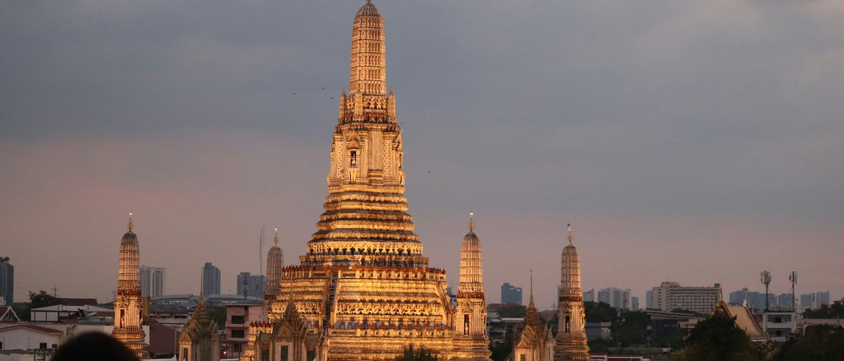 Wat Arun's golden spires lit by the last sunset light, with the Bangkok skyline blurring into pink twilight beyond
