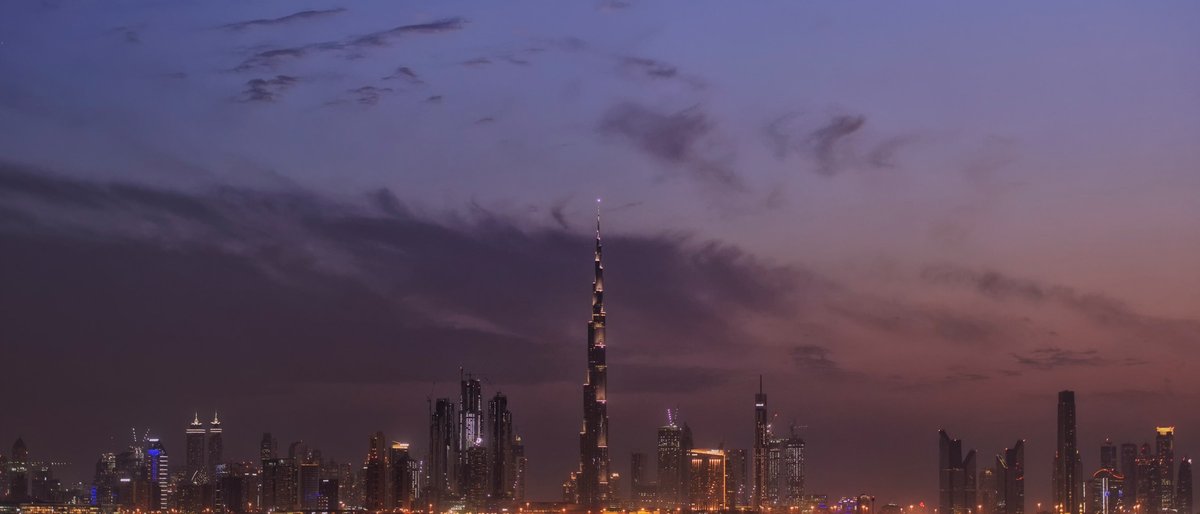 The Dubai skyline at violet twilight viewed across dark water, Burj Khalifa spearing high above the glittering Downtown and Business Bay towers while streaks of rose-mauve cloud drift over a deep indigo sky