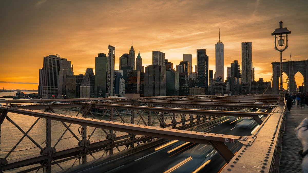Looking down the Brooklyn Bridge's steel trusses toward Lower Manhattan glowing under an amber sunset, traffic streaking into long-exposure light trails beside an antique lamppost on the wooden walkway