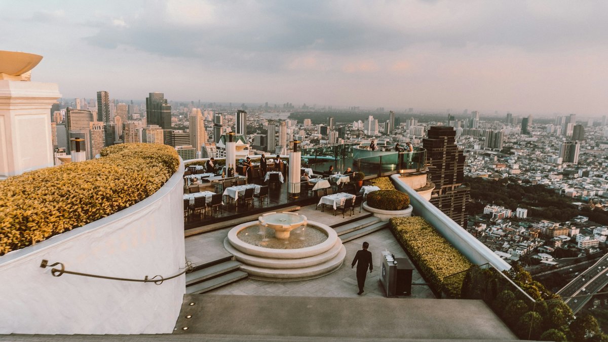 Bangkok's sky-high rooftop terrace at dusk, dressed tables and a stone fountain perched above the hazy sprawl of the city