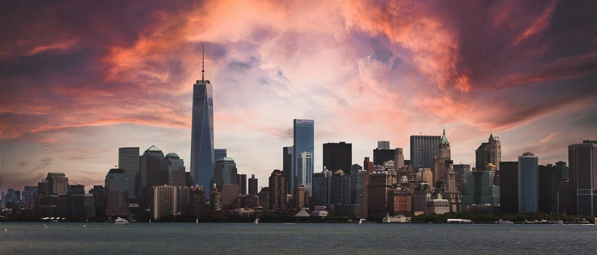 The Lower Manhattan skyline silhouetted across the Hudson with One World Trade Center spearing a sky of fiery pink and violet storm clouds at sunset, the harbor water dark and still in the foreground