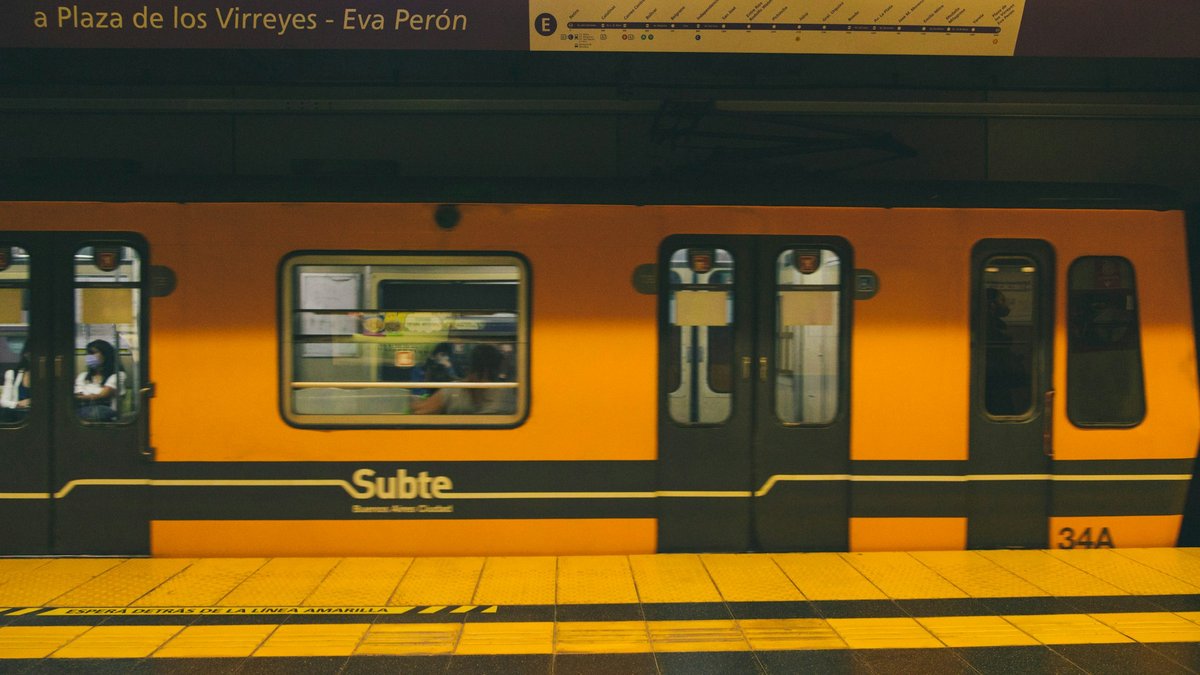 A yellow Subte Line E train stopped at the platform beneath a destination board reading 'Plaza de los Virreyes - Eva Perón', warm amber tunnel light washing the livery and safety stripe