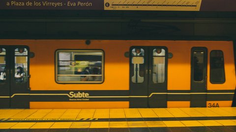 A yellow Subte Line E train stopped at the platform beneath a destination board reading 'Plaza de los Virreyes - Eva Perón', warm amber tunnel light washing the livery and safety stripe