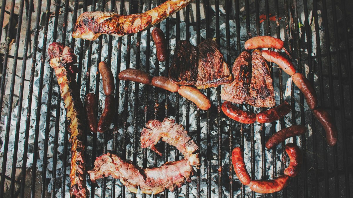 Overhead view of a South African braai mid-cook with coiled boerewors, lamb chops, ribs, sausages and seared steaks arranged across a charcoal-glowing cast-iron grill grate over white-ash embers