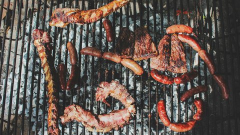 Overhead view of a South African braai mid-cook with coiled boerewors, lamb chops, ribs, sausages and seared steaks arranged across a charcoal-glowing cast-iron grill grate over white-ash embers