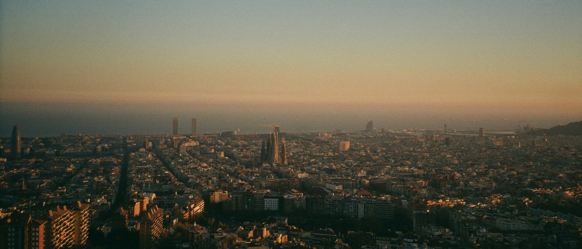 An aerial dusk panorama of Barcelona from the Bunkers del Carmel, the Sagrada Família and Torre Glòries rising above an endless grid of rooftops washed in molten gold