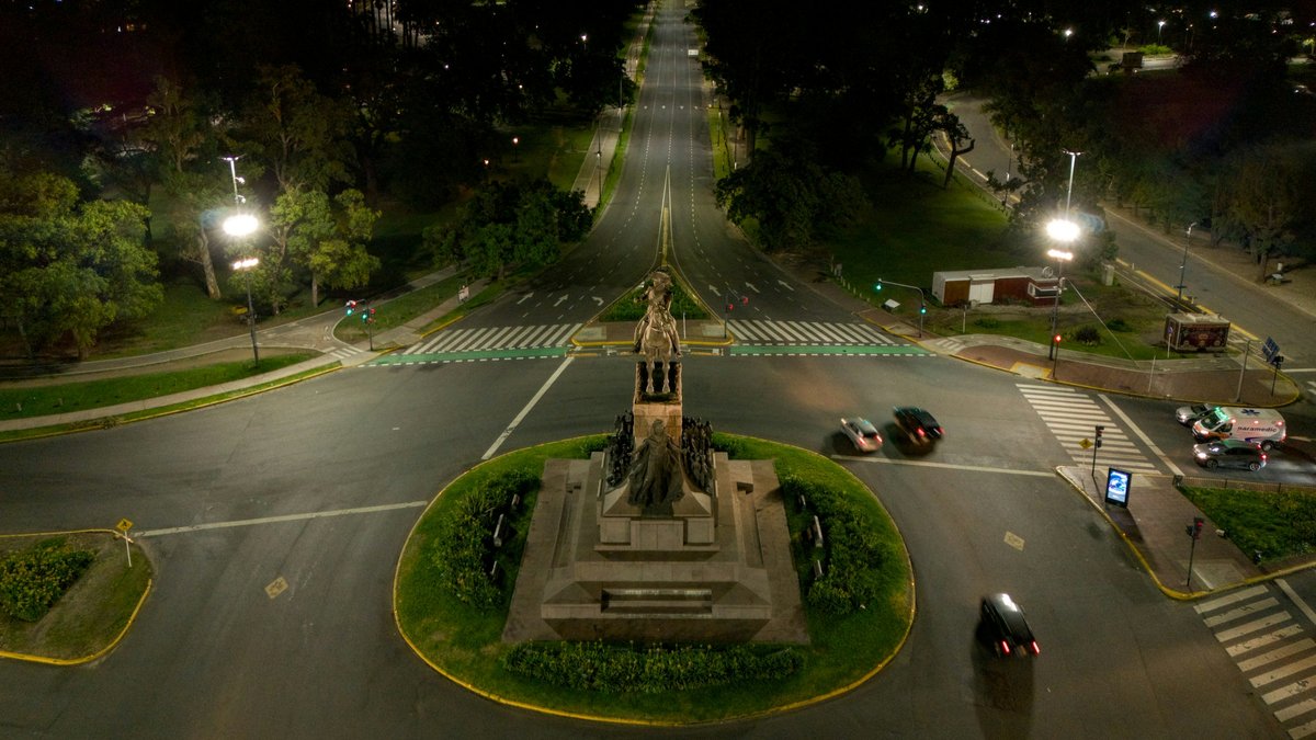 An aerial night view of a Buenos Aires avenue vanishing point through an equestrian-monument roundabout in Palermo, twin lamppost flares balancing the frame and traffic tracing thin red taillights across empty lanes