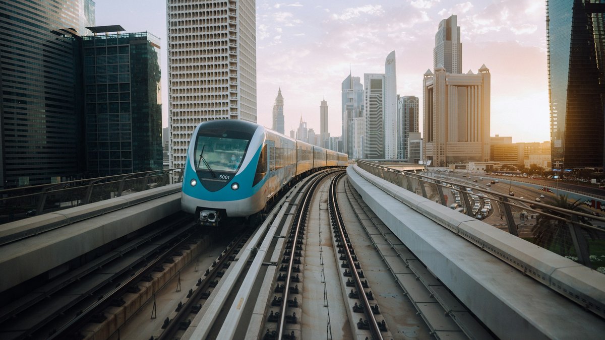 A blue-and-white Dubai Metro train curves along its elevated track between the Sheikh Zayed Road skyscrapers under a pastel late-afternoon sky, concrete sleepers and parallel rails vanishing toward the city