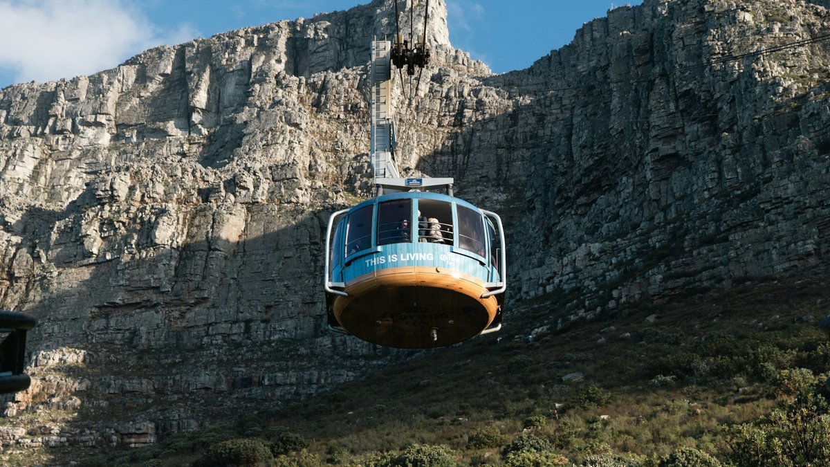 A teal Table Mountain Aerial Cableway rotair car descends mid-cable against the layered sandstone cliff face, fynbos scrub blanketing the lower slopes under a crisp blue sky