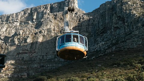 A teal Table Mountain Aerial Cableway rotair car descends mid-cable against the layered sandstone cliff face, fynbos scrub blanketing the lower slopes under a crisp blue sky