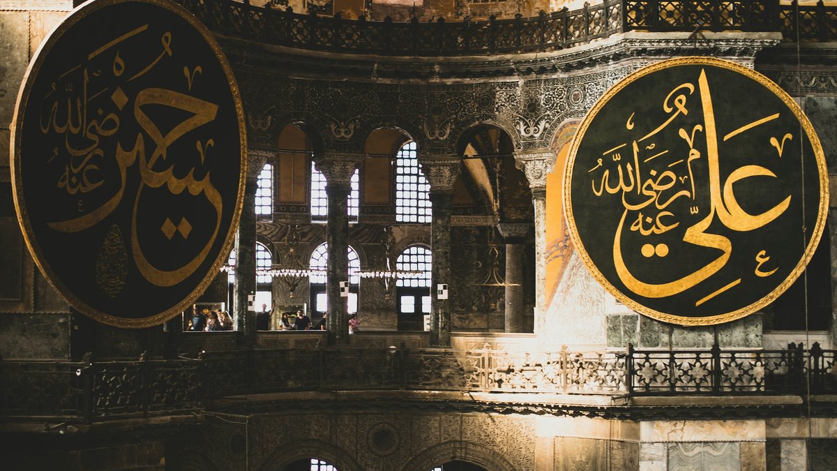 Two enormous black-and-gold calligraphy medallions flanking the nave of the Hagia Sophia, arched windows and Byzantine stonework glowing in soft interior light
