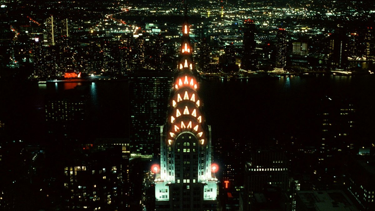 The Chrysler Building's stainless-steel art-deco crown glows in tiered orange chevrons against a sea of twinkling Midtown and Queens lights, the East River cutting a dark ribbon across the nighttime cityscape