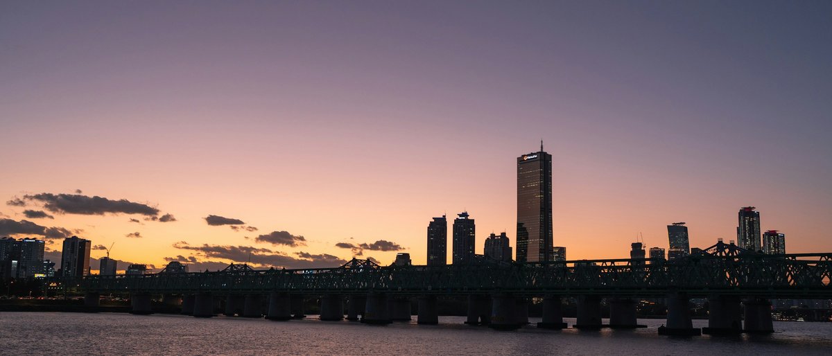 A railway bridge cuts across the Han River beneath Seoul's skyline at dusk, the 63 Building anchoring a horizon that melts from peach to deep violet as city lights flicker on across Yeouido