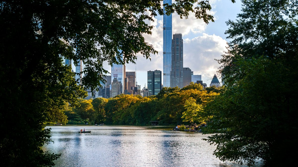 A wooden rowboat drifts on Central Park's Lake while the Billionaires' Row skyline — Central Park Tower and One57 — rises beyond a soft frame of late-summer foliage and lakeside trees