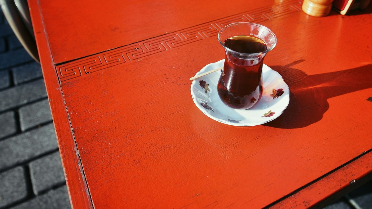 A tulip-shaped glass of Turkish çay on a bright red café table, casting a long afternoon shadow across the weathered stone beneath