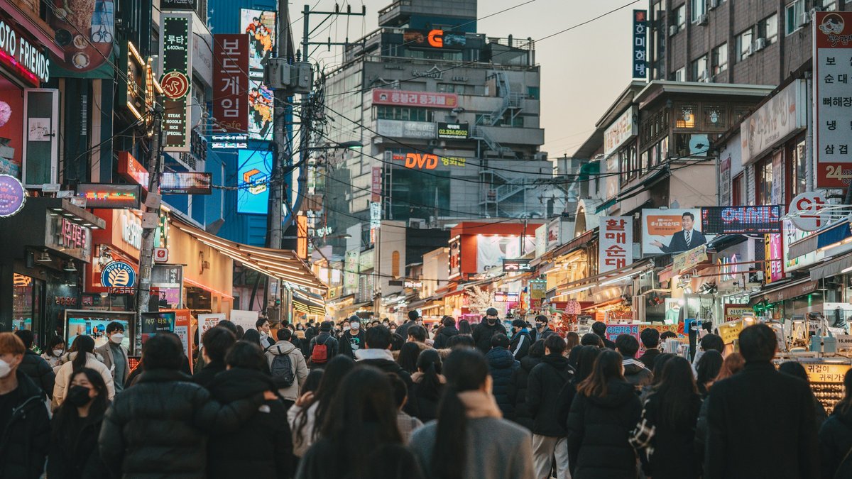 Crowds funnel down a Seoul backstreet at blue hour, puffer-jacketed silhouettes threading between stacks of Hangul neon signs, tangled overhead wires and warm shop-window glow