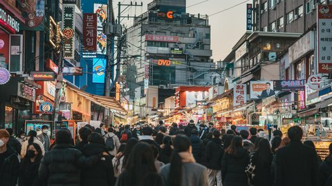 Crowds funnel down a Seoul backstreet at blue hour, puffer-jacketed silhouettes threading between stacks of Hangul neon signs, tangled overhead wires and warm shop-window glow