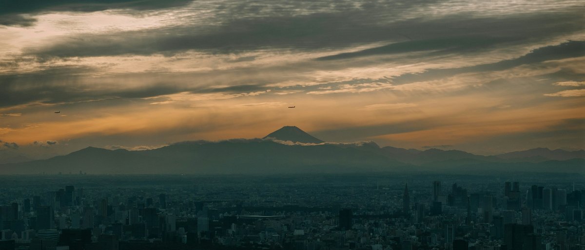 Mount Fuji's dark silhouette floats above Tokyo's endless grid of towers at dusk, the sky melting from peach to indigo as the city's lights begin to flicker on